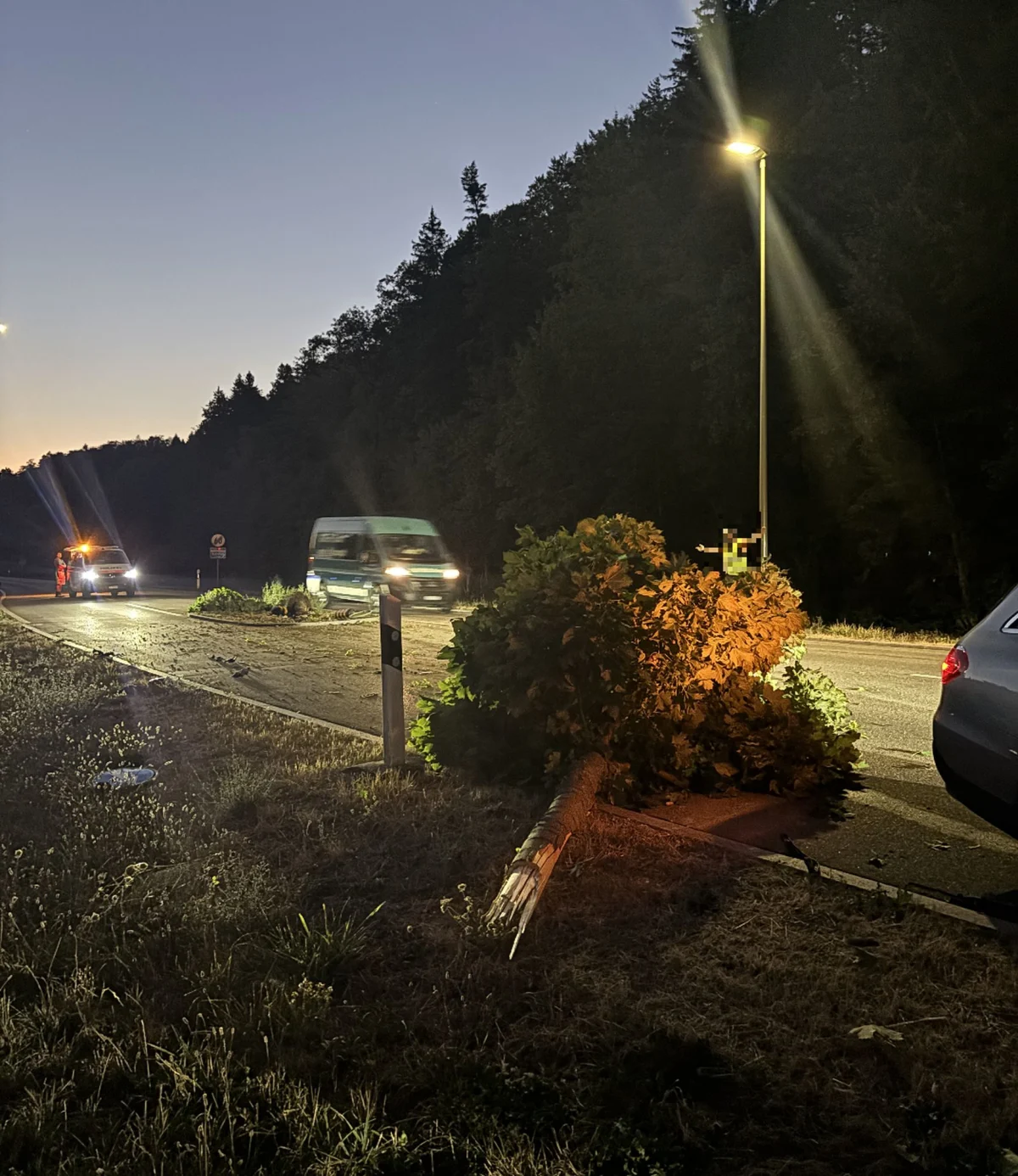 Ein Baum liegt quer auf einer Landstraße, beleuchtet von einer Straßenlaterne, während Fahrzeuge in der Dämmerung vorbeifahren.