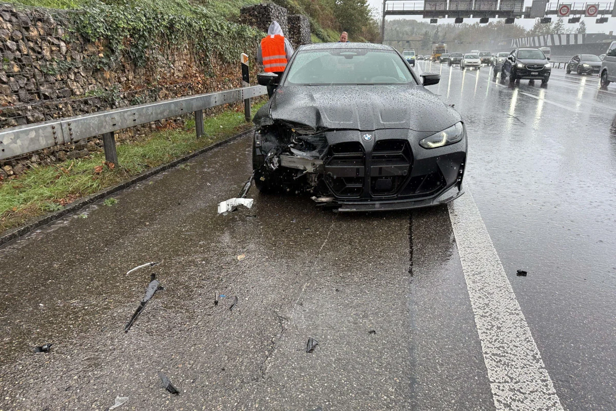 Ein schwarzes Auto, dessen Vorderteil beschädigt ist, steht auf einer regennassen Autobahnspur. Trümmerteile umgeben das Fahrzeug.