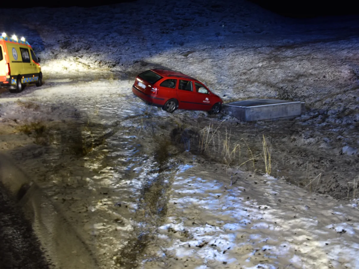 Ein rotes Auto steckt seitlich in einem schneebedeckten Graben fest. Ein Ambulanzfahrzeug steht in der Nähe, Licht strahlt auf die Szene.