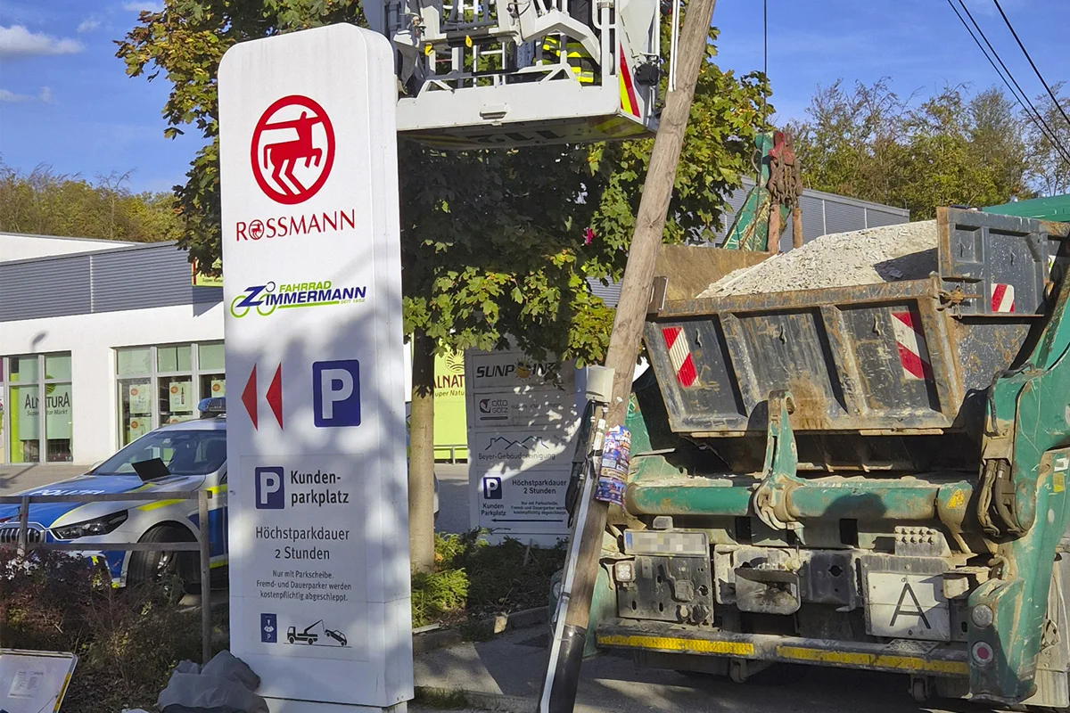 Ein Baustellenfahrzeug mit Kies beladen steht neben einem Geschäftsschild, darunter ein Polizeifahrzeug auf einem Parkplatz.