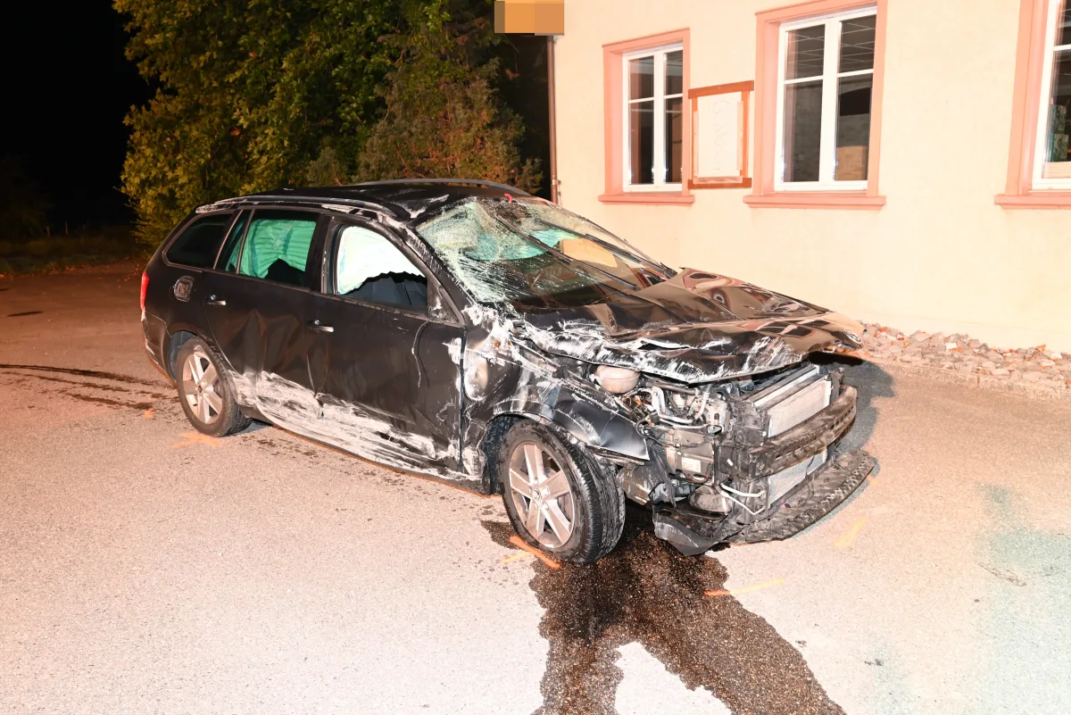 Ein schwer beschädigtes Auto steht vor einem Gebäude, die Vorderseite ist stark eingedellt und Scheiben sind beschädigt.