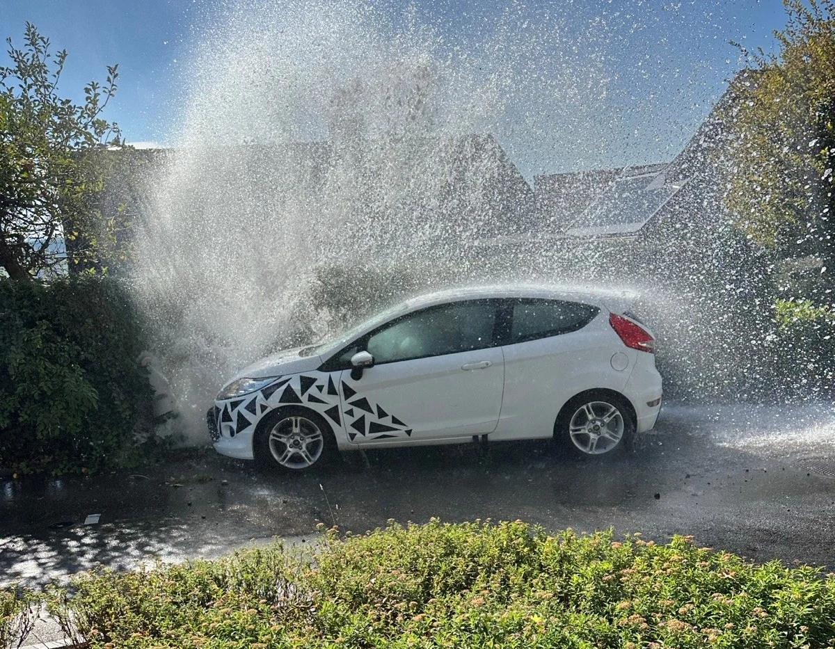 Ein weißes Auto ist mit einem Hydranten kollidiert. Wasser schießt hoch in die Luft, während das Fahrzeug im Vordergrund teilweise sichtbar ist.