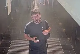Ein Mann in dunkler Sportkleidung steht in einem gefliesten Korridor, Blick zur Kamera, in einer hell erleuchteten Umgebung.