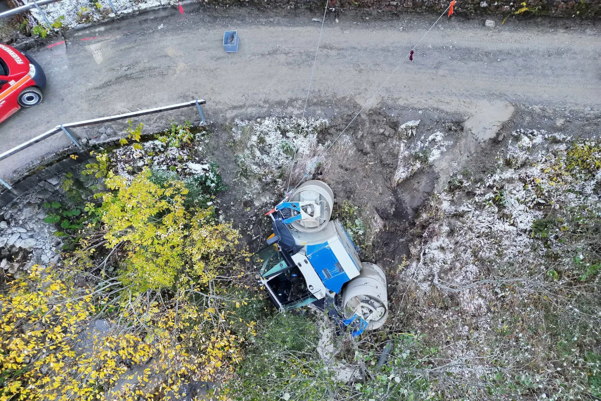 Ein umgestürztes Baugerät, möglicherweise ein Betonmischer, liegt in einem Graben neben einer kurvigen Straße, umgeben von Herbstlaub.