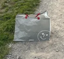 Eine graue Papiertüte mit roten Henkeln und einem Smiley liegt auf einem Schotterweg neben grünen Gräsern.