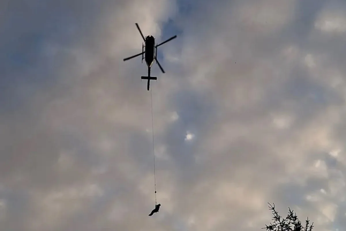 Ein Helikopter schwebt am Himmel mit einer Person, die an einem Seil hängt, bei bewölktem Wetter; unten sind Baumspitzen sichtbar.