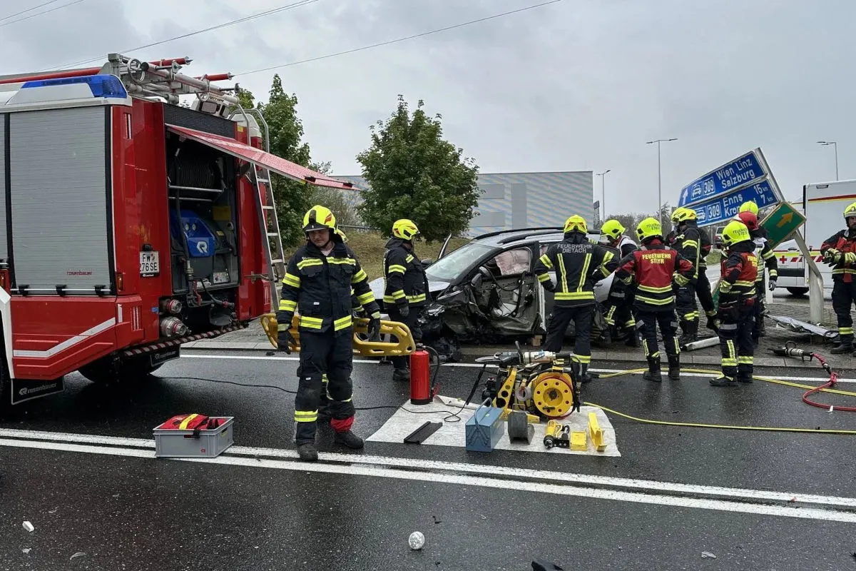 Feuerwehrleute in voller Ausrüstung stehen um ein stark beschädigtes Auto, nahe eines roten Einsatzfahrzeugs. Rettungswerkzeuge sind auf der Straße verteilt.