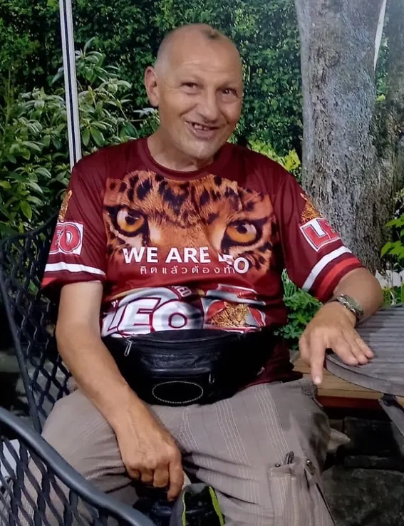 Ein Mann sitzt draußen auf einem Stuhl vor einem grünen Hintergrund. Er trägt ein auffälliges T-Shirt und lächelt in die Kamera.