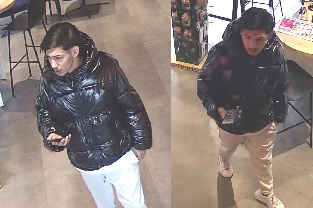 Ein Mann mit dunklen Haaren trägt eine schwarze, glänzende Jacke und weiße Hose. Er hält ein Handy und befindet sich in einem Geschäft.