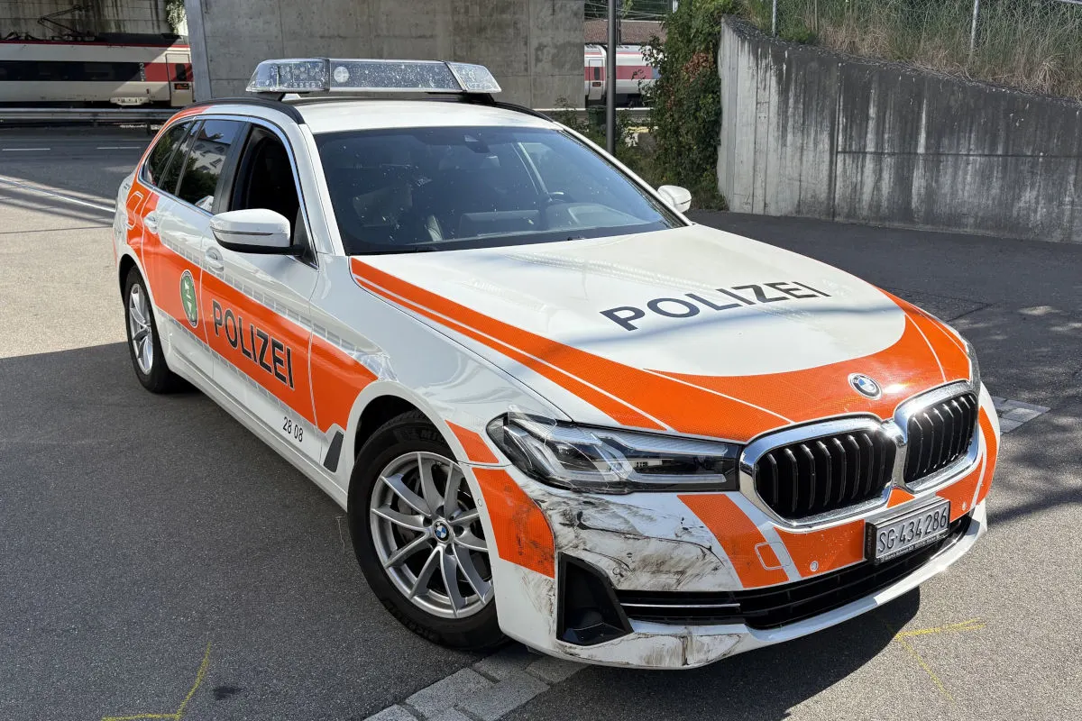 Ein weiß-orangefarbenes Polizeiauto zeigt Schäden und Schmutz an der Vorderseite. Im Hintergrund ist ein Zug zu sehen.