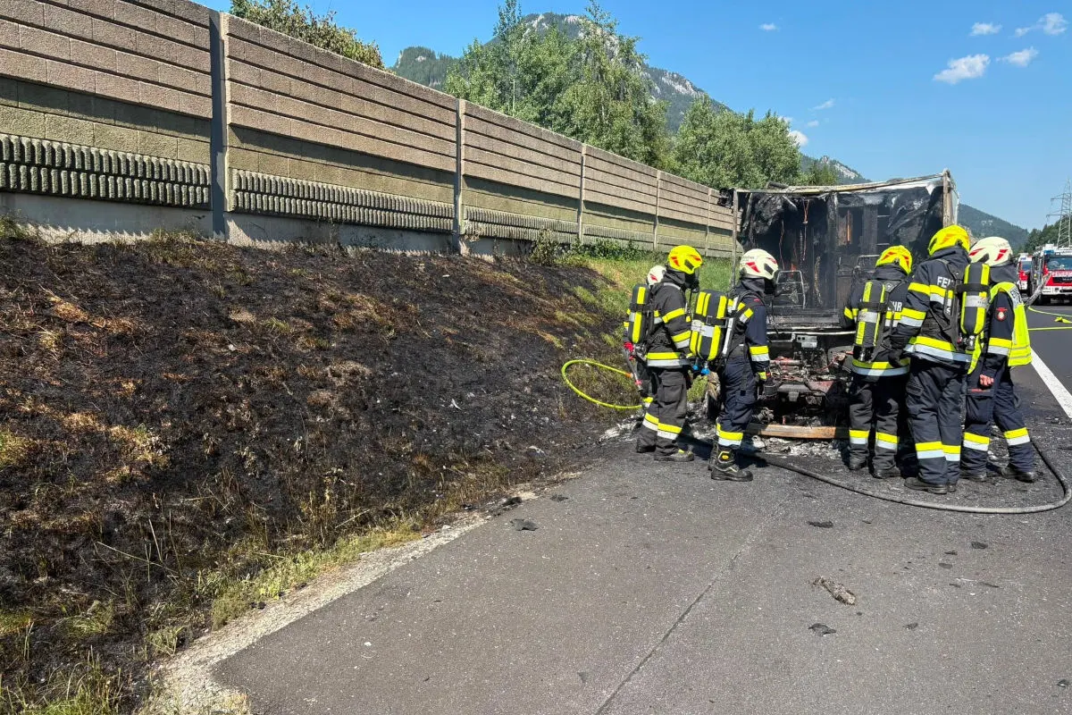 Mehrere Feuerwehrleute in Schutzkleidung stehen um die Überreste eines ausgebrannten Fahrzeugs auf einer Autobahn herum. Im Hintergrund ist eine Betonlärmschutzwand zu sehen.