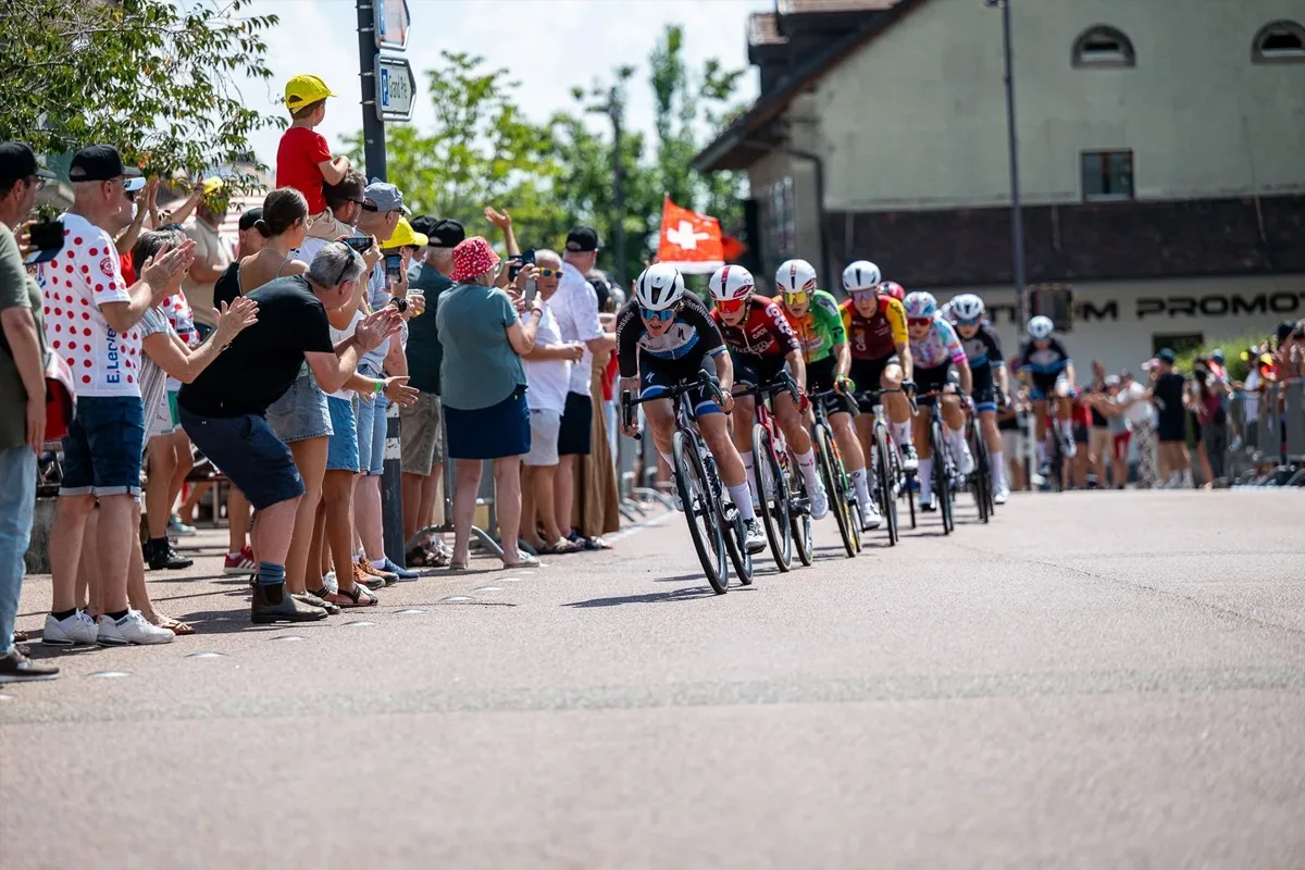 Eine Gruppe von Radfahrern fährt in einem Rennen, während Zuschauer am Straßenrand begeistert applaudieren. Eine Schweizer Flagge ist sichtbar.