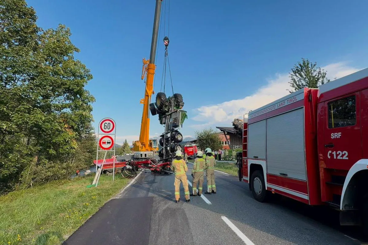 Ein Kran hebt einen umgekippten Traktor neben einem Feuerwehrfahrzeug auf einer Straße auf. Drei Feuerwehrleute beobachten die Aktion.