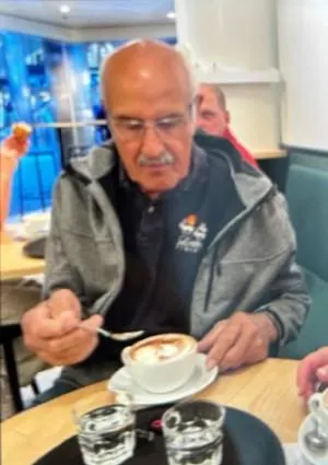 Ein älterer Mann sitzt an einem Holztisch in einem belebten Café und hält eine Tasse Cappuccino in der Hand.