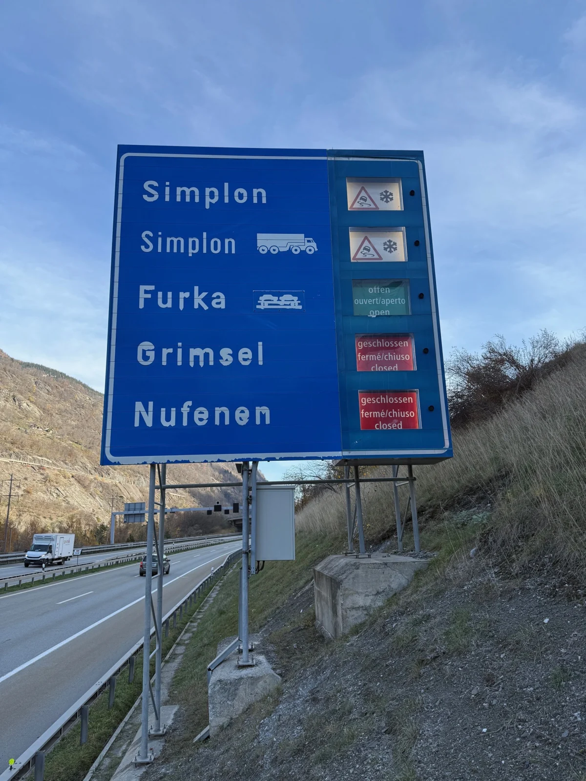 Ein großes Verkehrszeichen zeigt Informationen zur Öffnung und Schließung der Pässe Simplon, Furka, Grimsel und Nufenen. Die Anzeige weist auf winterliche Bedingungen hin.