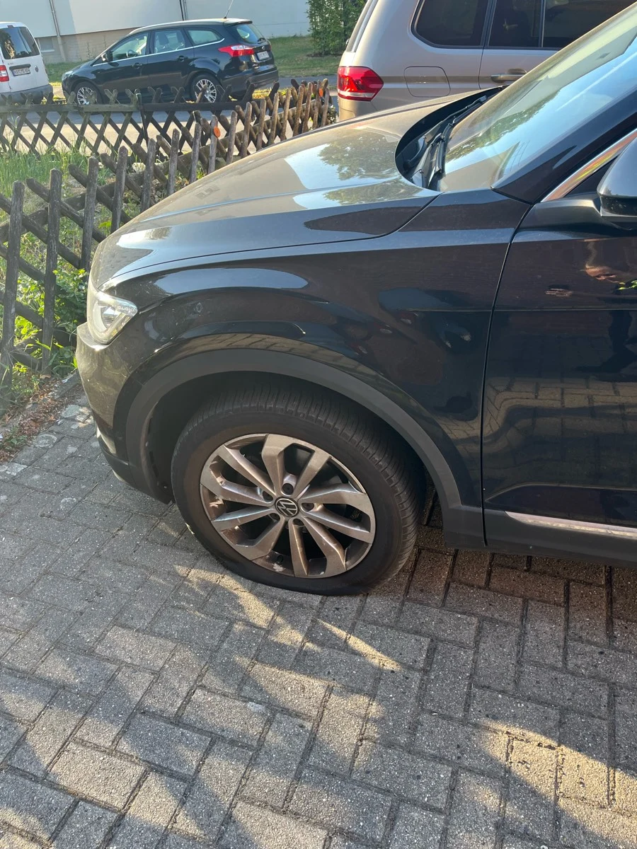 Ein dunkler VW steht auf einem Pflastersteinweg, der vordere linke Reifen ist platt. Im Hintergrund sind zwei weitere Autos geparkt.