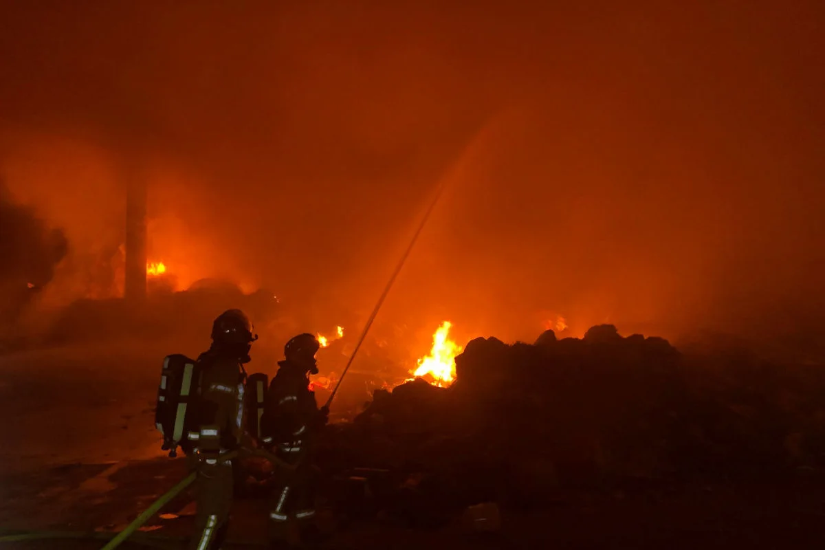 Im Bild sind zwei Feuerwehrleute zu sehen, die mit einem Schlauch versuchen, ein großes Feuer zu löschen. Rauch und Flammen dominieren die Szenerie.