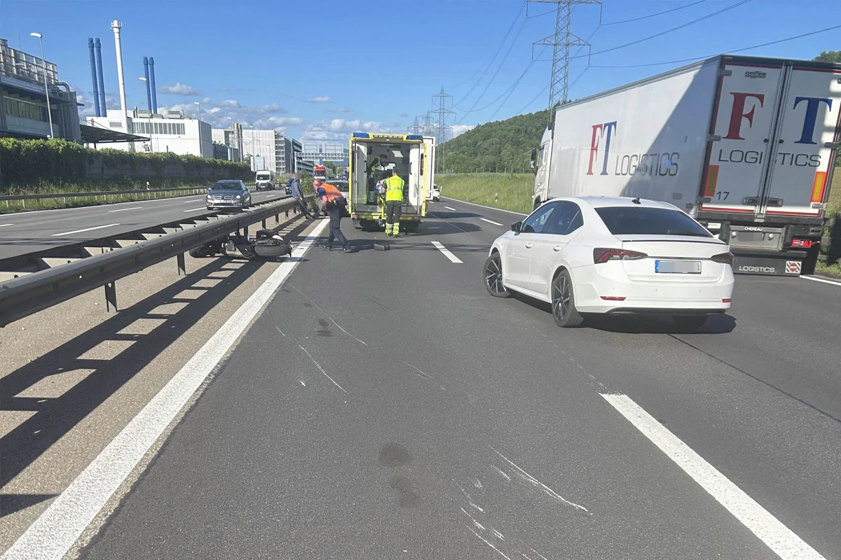 Auf einer Autobahn hat sich ein Unfall ereignet. Ein Motorrad liegt neben der Leitplanke, und Rettungskräfte stehen bei einem Ambulanzfahrzeug. Fahrzeuge sind auf der Fahrbahn zu sehen.