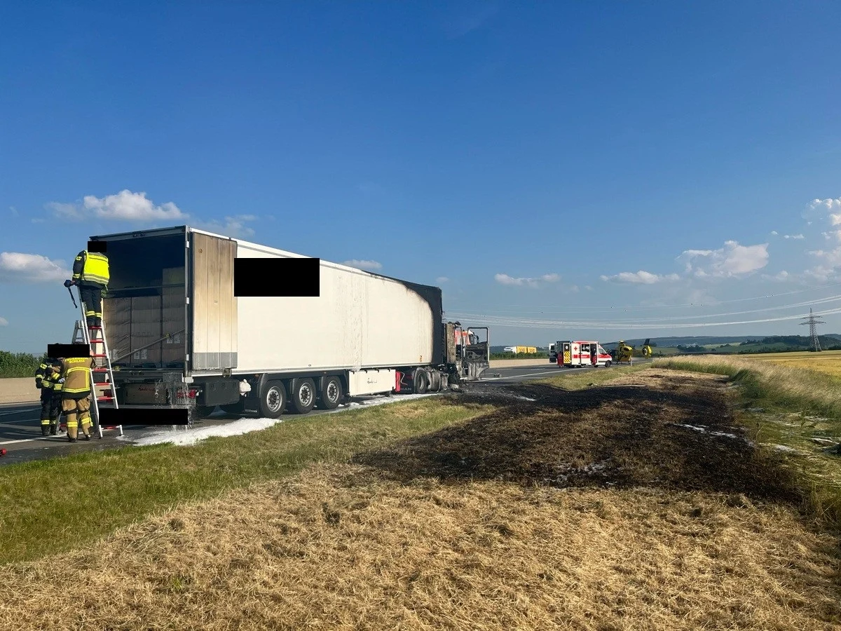 Ein ausgebrannter Lkw befindet sich auf einer Straße, umgeben von Rettungskräften und Fahrzeugen. Der Himmel ist klar und die Straße ist trocken.