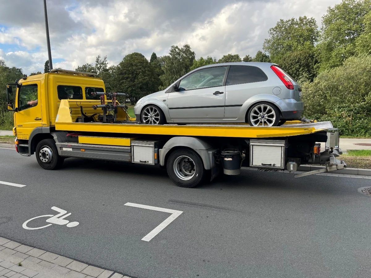 Ein silberfarbenes Auto steht auf einem gelben Abschleppwagen, der auf einer Straße parkt. Im Hintergrund sind Bäume und Wolken zu sehen.