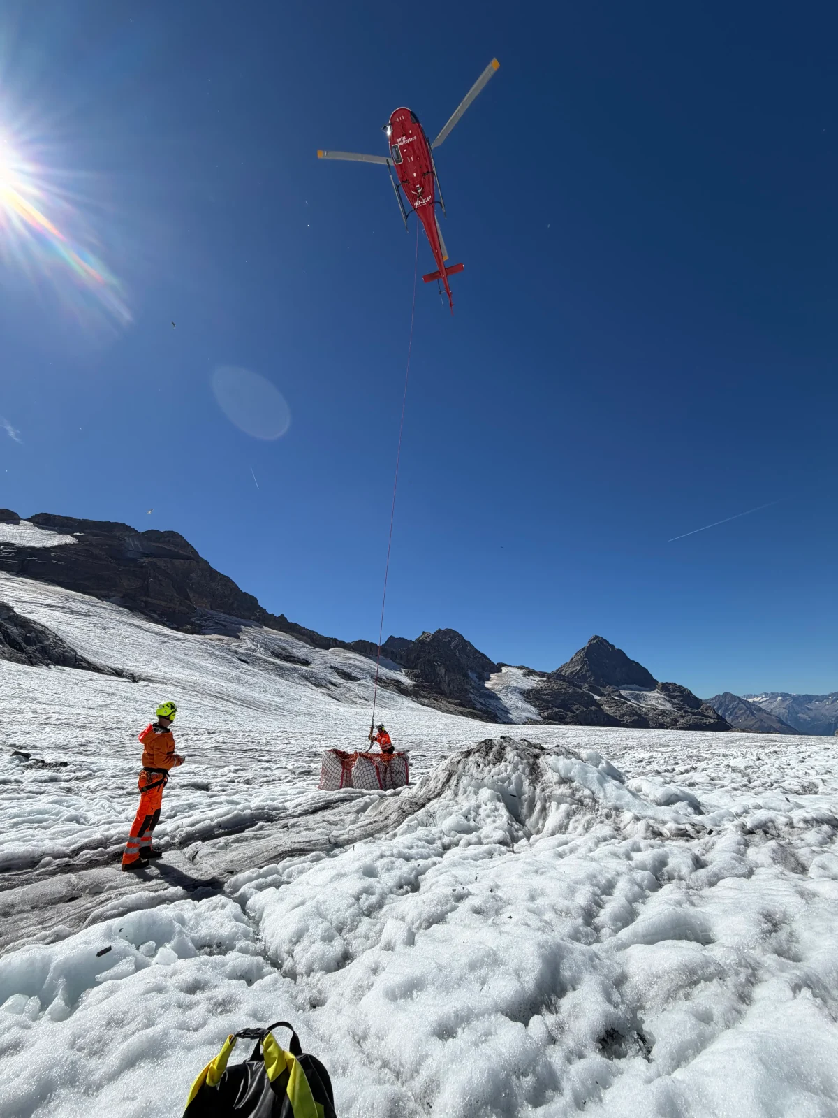 Ein roter Helikopter fliegt über eine schneebedeckte Gletscherlandschaft. Zwei Arbeiter in orangenen Schutzanzügen stehen auf dem Eis und sichern eine Frachtkiste.