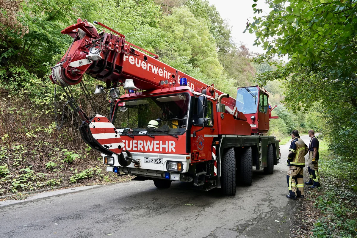 Ein großer, roter Feuerwehrkran steht auf einer asphaltierten Straße, umgeben von dichtem grünen Wald. Zwei Feuerwehrleute in Uniform stehen daneben.