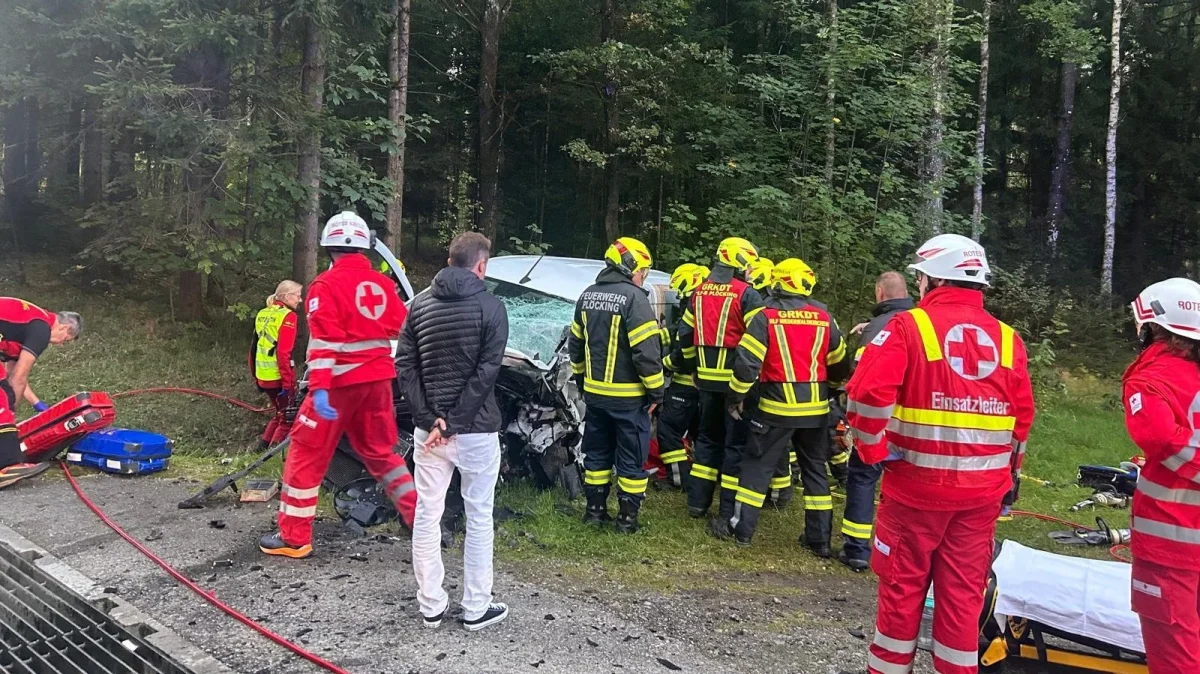 Mehrere Feuerwehrleute und Rettungskräfte stehen um ein beschädigtes Auto in einem Waldgebiet. Ein Mann in zivil beobachtet die Szene.