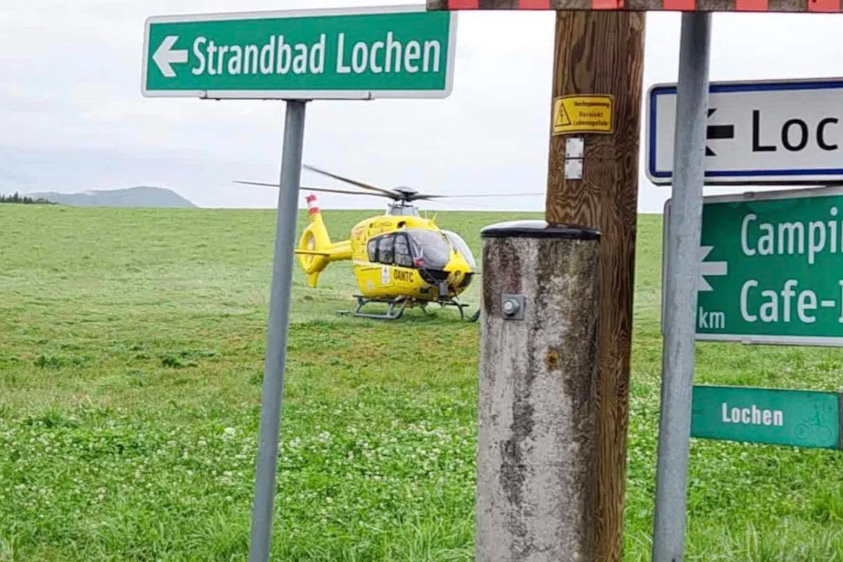 Ein gelber Rettungshubschrauber steht auf einer grünen Wiese hinter Wegweisern, die zum Strandbad Lochen führen.