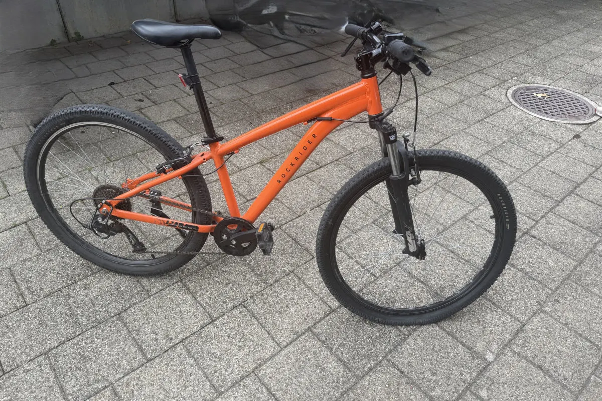 Ein orangefarbenes Mountainbike steht seitlich auf einem gepflasterten Gehweg. Es zeigt deutliche Gebrauchsspuren.