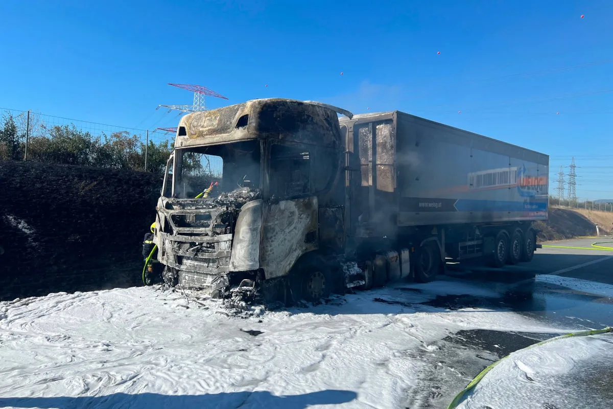 Ein Lkw mit stark verbrannter Front steht auf einer Straße, umgeben von Löschschaum. Der Himmel ist klar und blau.