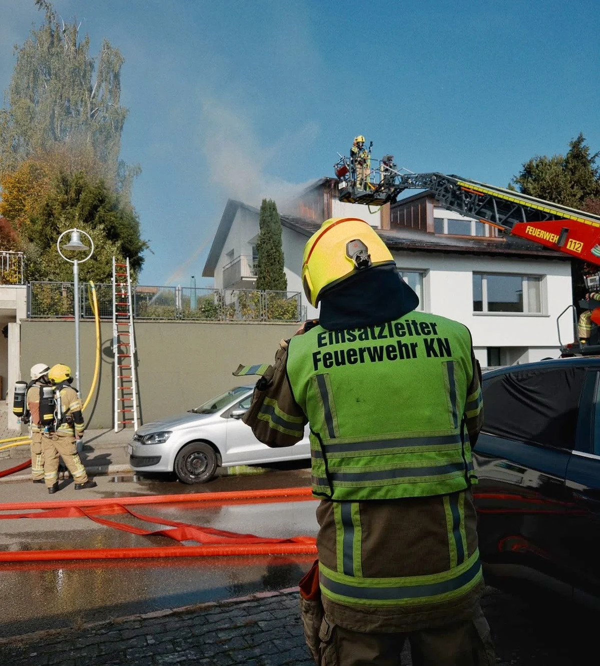 Mehrere Feuerwehrleute arbeiten an einem Wohnhausbrand. Eine Drehleiter ist im Einsatz, und Schläuche liegen auf der Straße.