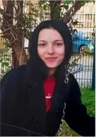 Eine junge Frau in einem schwarzen Mantel sitzt outdoor vor einem Baum. Im Hintergrund ist ein Zaun erkennbar.