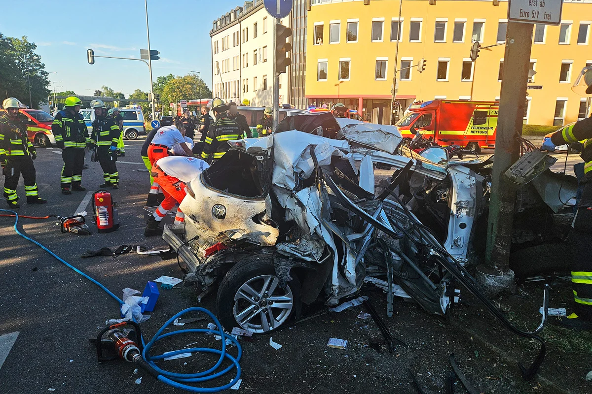 Ein stark beschädigtes Auto steht an einer Kreuzung, umgeben von Feuerwehrleuten und Sanitätern. Rettungsfahrzeuge und Ausrüstung sind vor Ort sichtbar.