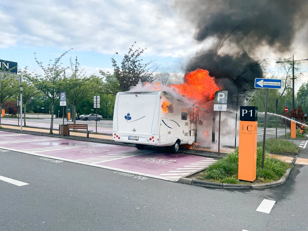 Auf einem Parkplatz steht ein Wohnmobil in Flammen. Dicker Rauch steigt empor, während das Fahrzeug im oberen Bereich stark beschädigt ist.