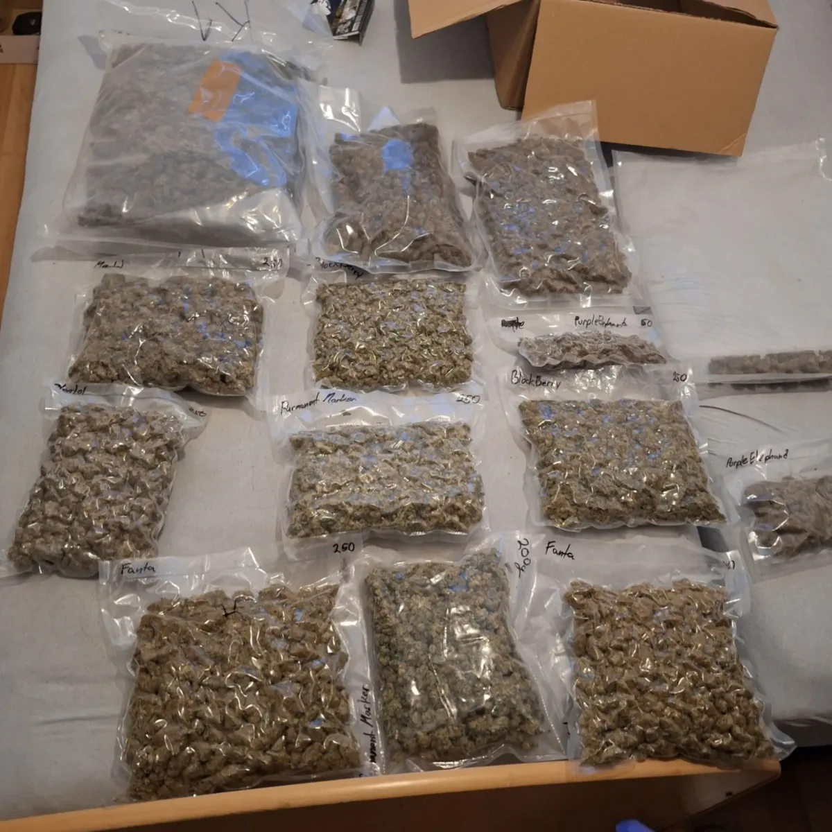 Auf einem Tisch liegen viele vakuumverpackte Plastiktüten mit Cannabis, teils beschriftet. Im Hintergrund steht ein offener Karton.