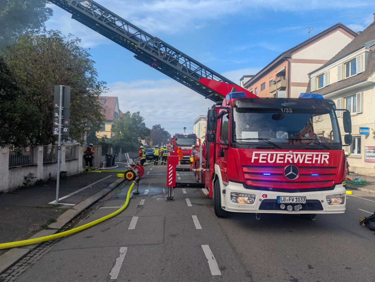 Ein rotes Feuerwehrfahrzeug mit einer ausgefahrenen Drehleiter steht auf einer Straße. Feuerwehrleute sind im Hintergrund im Einsatz.