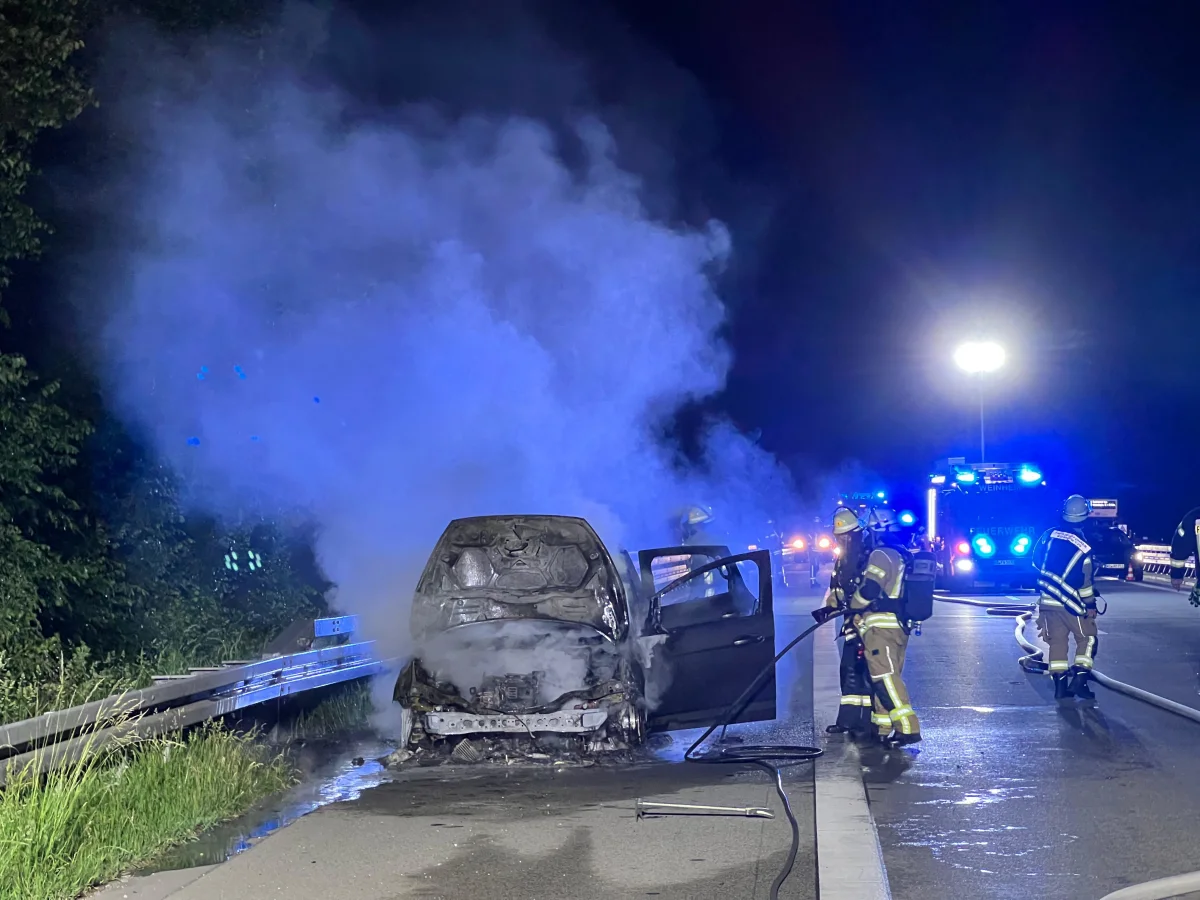 Auf einer Autobahn steht ein ausgebranntes Auto, umgeben von Rauch, während Feuerwehrleute mit Löscharbeiten beschäftigt sind. Im Hintergrund sind Einsatzfahrzeuge mit Blaulicht zu sehen.