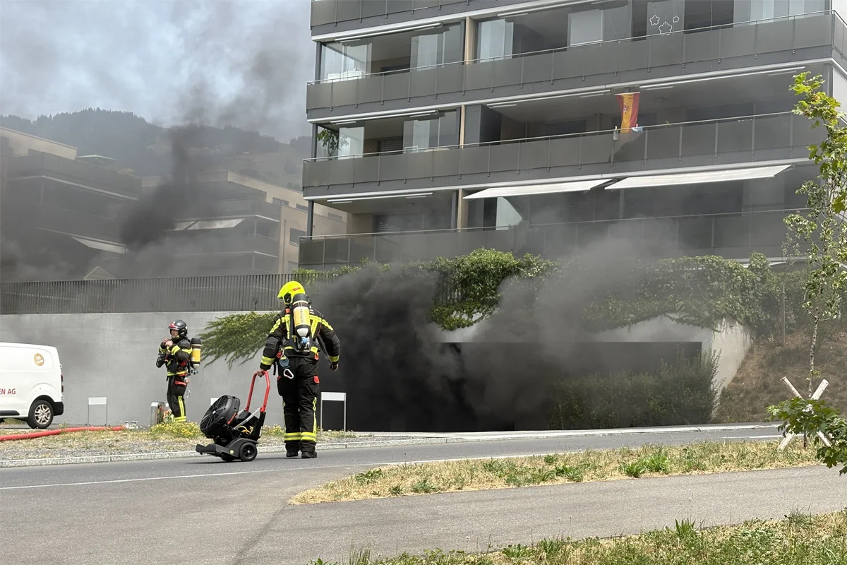 Zwei Feuerwehrleute in Schutzkleidung stehen vor einem rauchenden Gebäude, schwarzer Rauch steigt aus einer Tiefgarage auf.