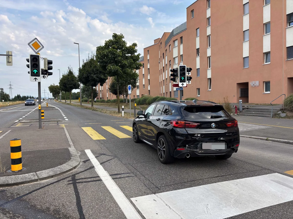 Ein schwarzer BMW X2 steht an einer städtischen Kreuzung mit Ampeln, während im Hintergrund ein Wohngebäude zu sehen ist.