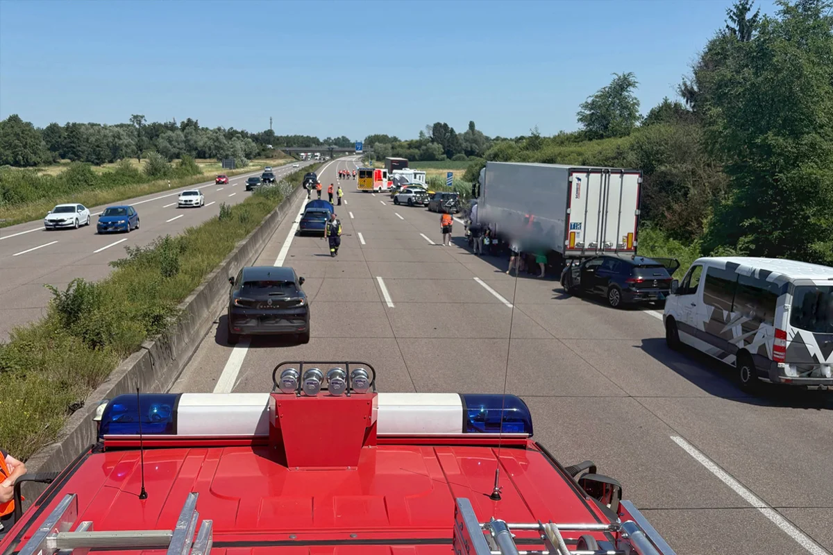 Auf einer Autobahn stehen Fahrzeuge, darunter ein großer Lkw und Einsatzfahrzeuge. Personen sind zu Fuß unterwegs.
