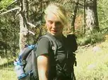 Eine Person mit hellem Haar und dunklem Rucksack wandert in einem Wald mit Bäumen und Gras.