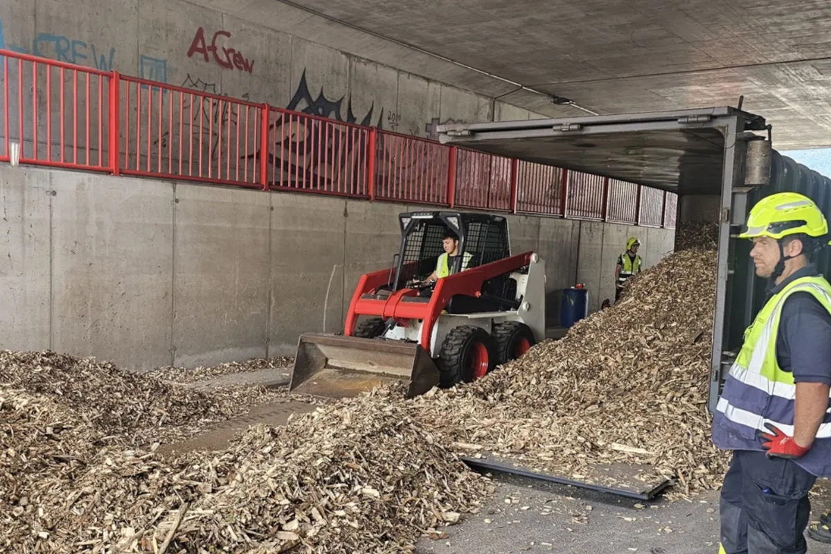 Ein Bobcat-Bagger wird eingesetzt, um Holzschnitzel unter einer Betonbrücke zu beseitigen. Arbeiter mit Schutzkleidung sind vor Ort.