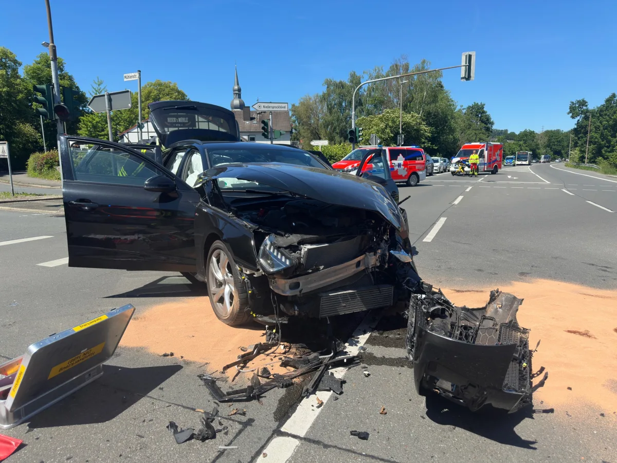 Ein schwarzes Auto mit stark beschädigter Front an einer Kreuzung. Rettungsfahrzeuge und Sanitäter sind im Hintergrund zu sehen.