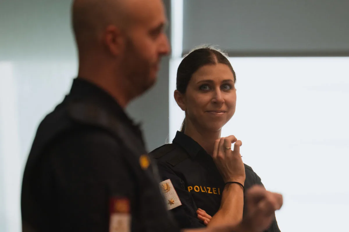 Eine Polizistin in Uniform lächelt in die Kamera, während ein Kollege unscharf im Vordergrund steht.