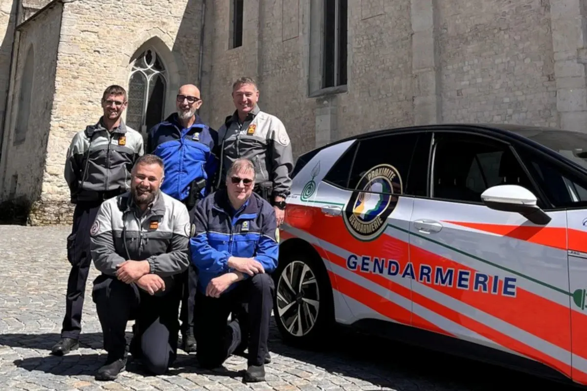 Fünf Männer in blauen und grauen Uniformen posieren neben einem Auto mit der Aufschrift 'Gendarmerie' vor einem Steingebäude. Das Fahrzeug hat auffällige rote und grüne Streifen.