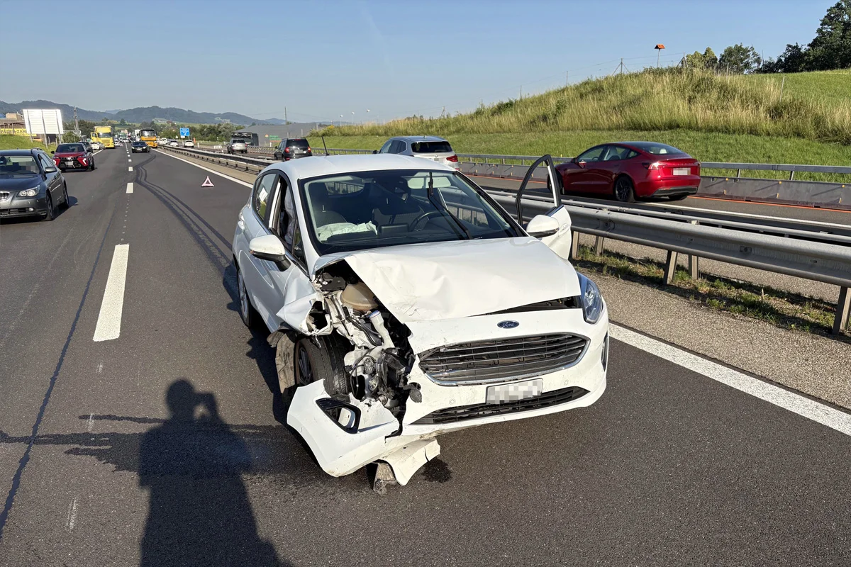 Ein weißes Auto mit stark beschädigtem Vorderteil steht auf einer Autobahn, umgeben von mehreren anderen Fahrzeugen. Die Fahrertür ist geöffnet.