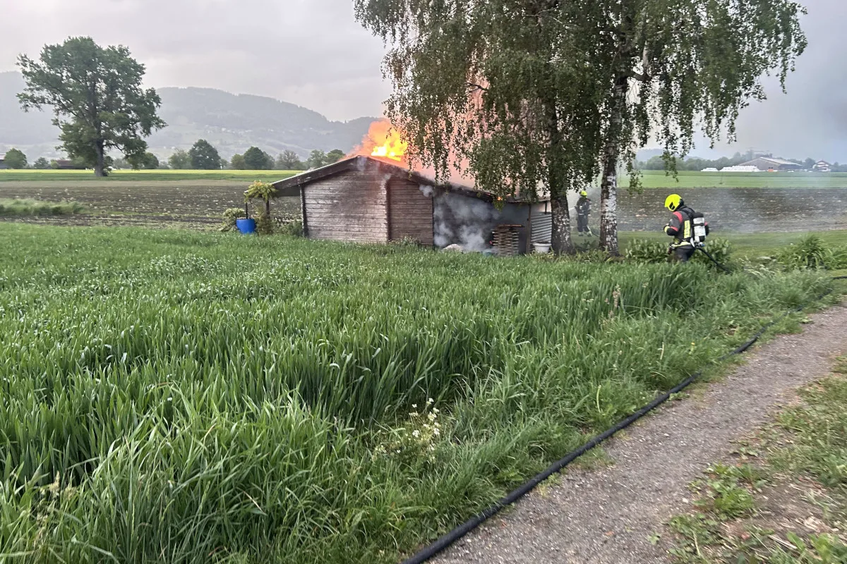 Ein großes Feuer lodert im Dach eines Holzgebäudes auf einem Feld. Zwei Feuerwehrleute mit Ausrüstung stehen neben dem Gebäude und versuchen, das Feuer zu löschen.