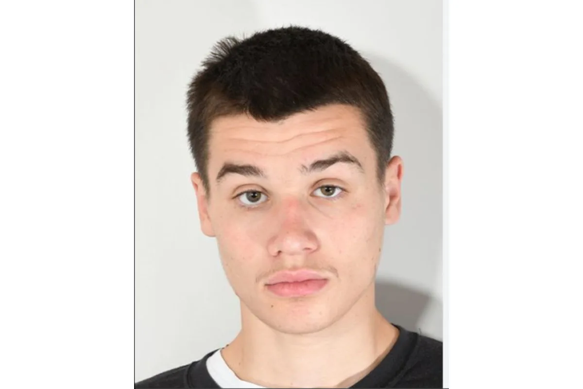 Das Bild zeigt einen jungen Mann mit dunklem, kurzem Haar vor einem neutralen Hintergrund. Er blickt direkt in die Kamera und trägt ein dunkles Oberteil.