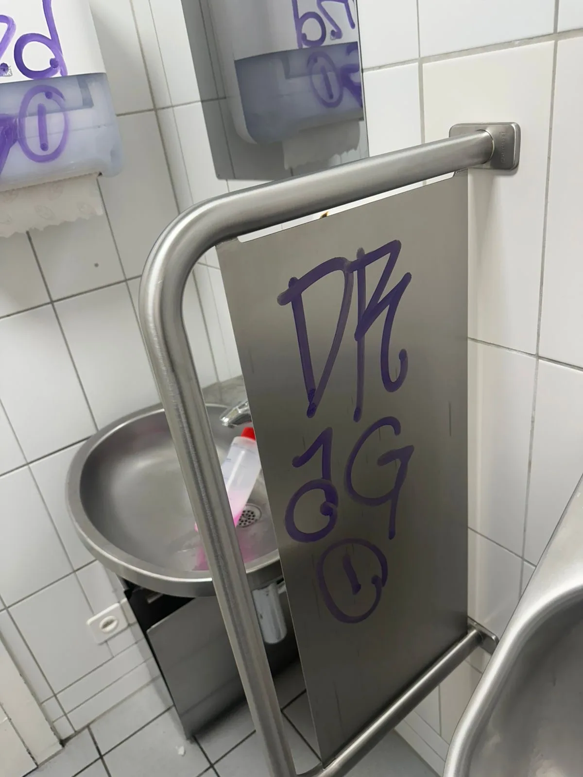 In einer öffentlichen Toilette ist auf einer Metallfläche lila Graffiti zu sehen, daneben befinden sich ein Waschbecken und Papierhandtuchspender.