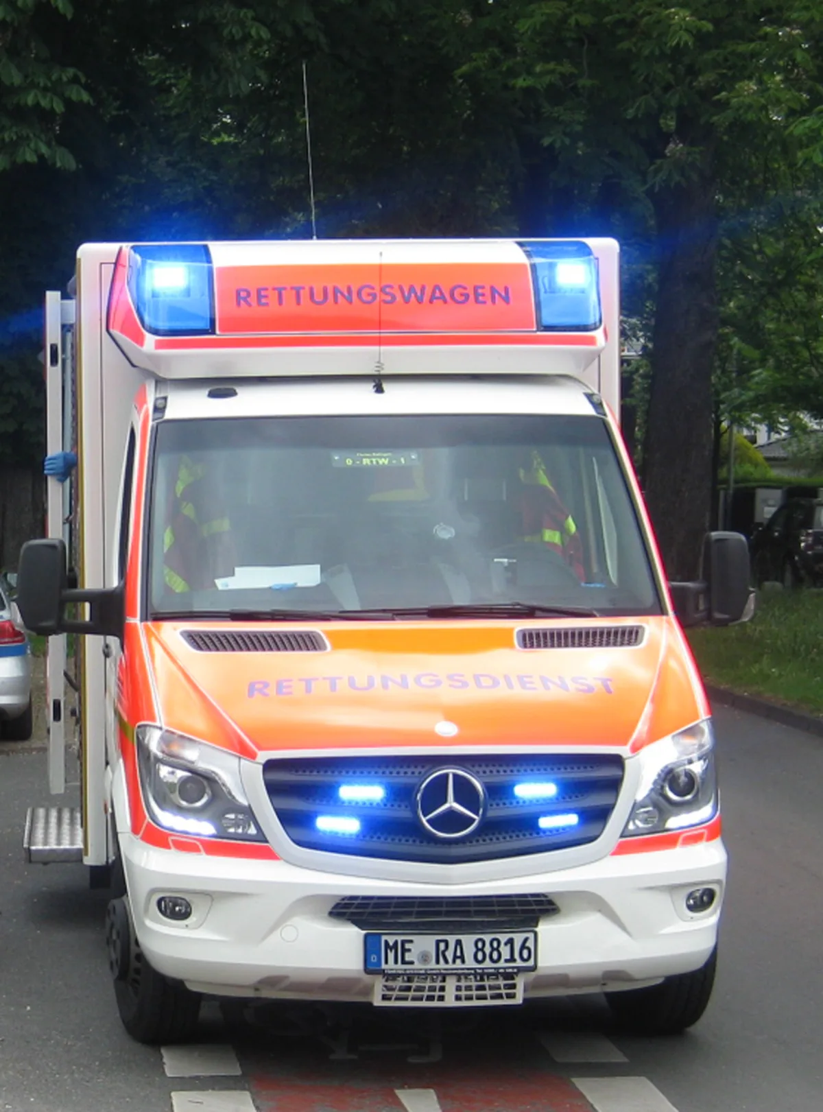 Ein Rettungswagen der Marke Mercedes-Benz in städtischem Umfeld mit aktiviertem Blaulicht. Der Aufbau ist orange-weiß, mit der Aufschrift 'RETTUNGSDIENST'.